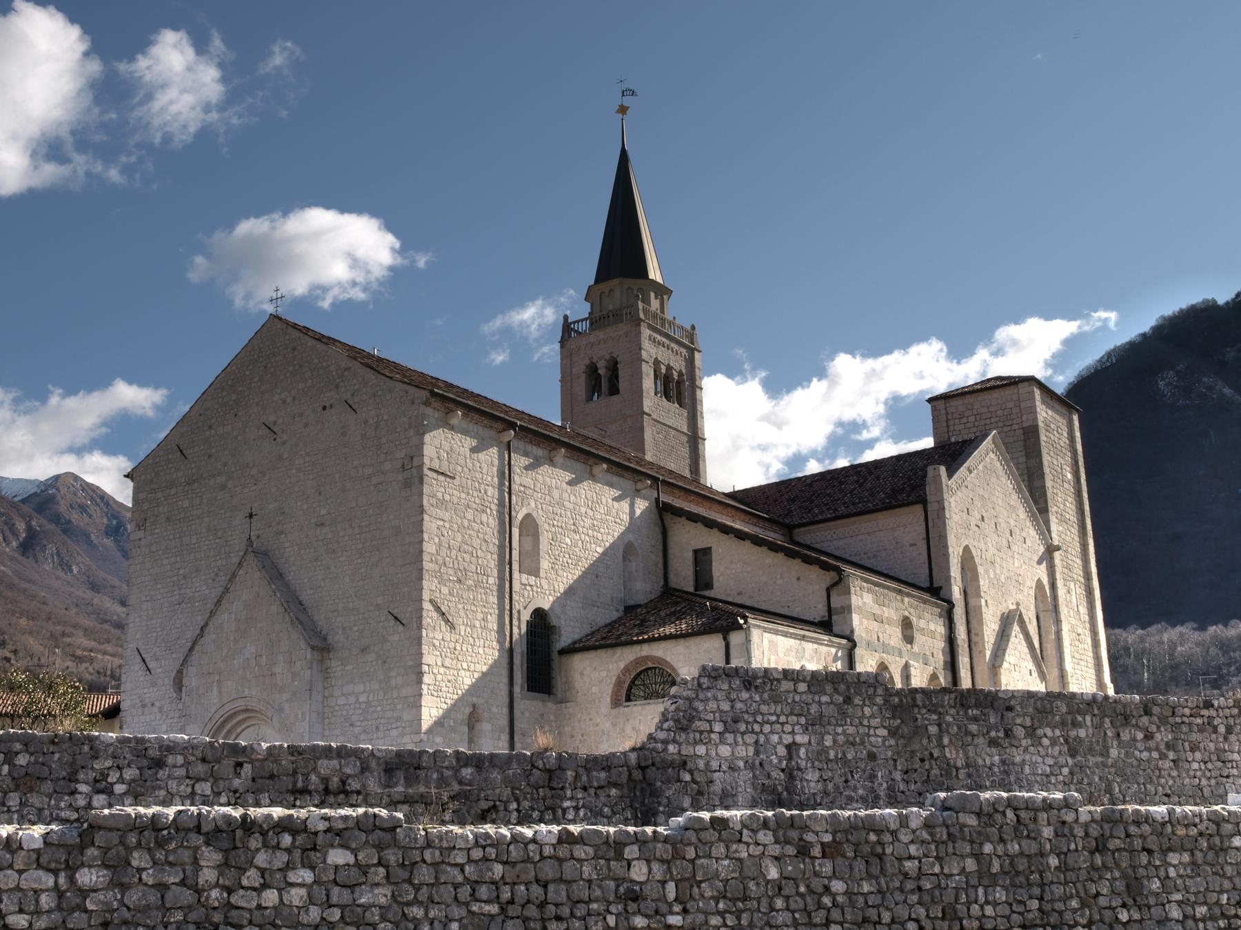 Venzone cathedral and bell tower behind medieval stone walls under blue sky